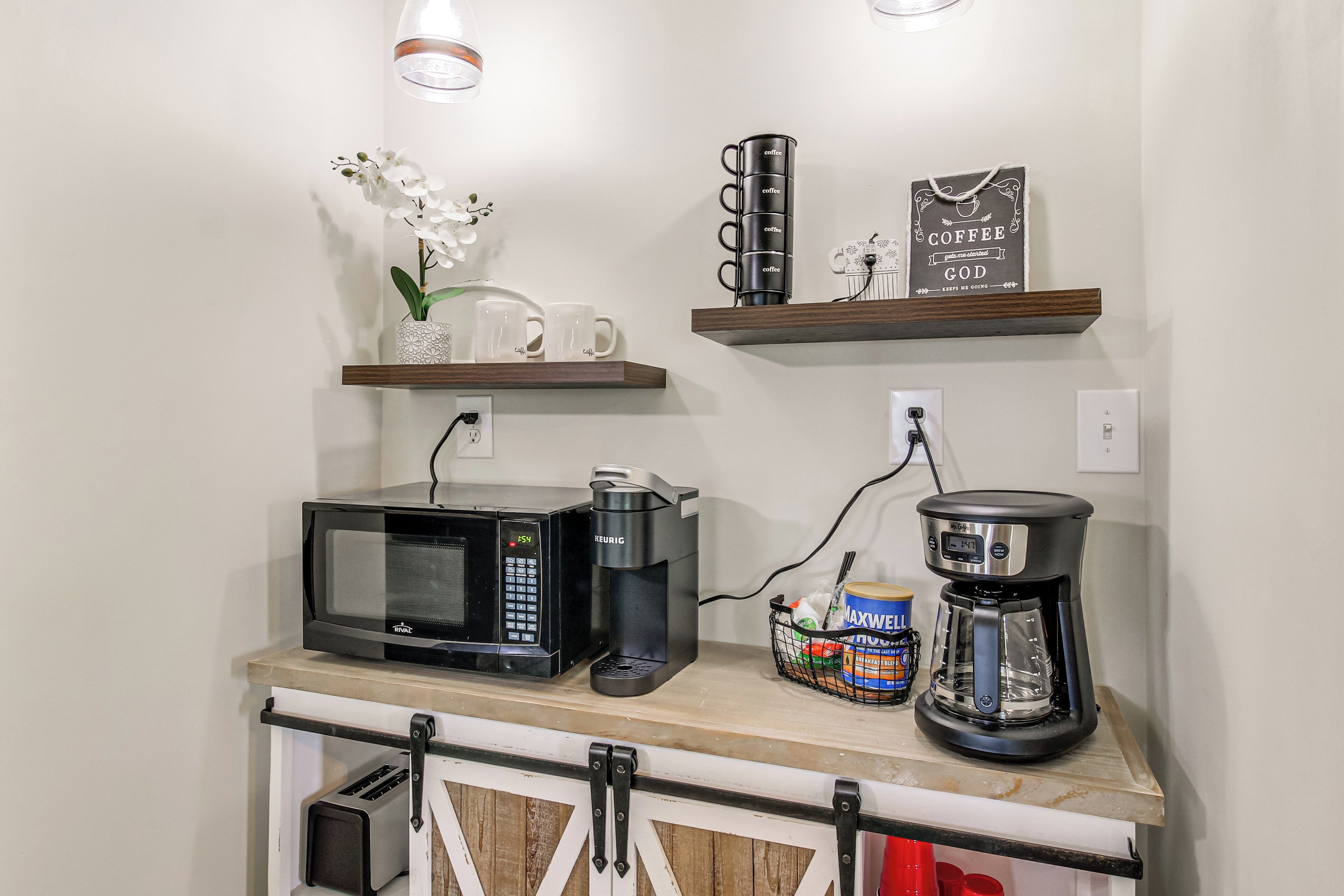 Une petite cuisine équipée d’un four à micro-ondes, d’une machine à café et d’un grille-pain. On peut y voir des fleurs blanches, une tasse à café et un panneau décoratif sur le thème du café, le tout placé sur les étagères.