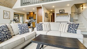 A spacious living area with a high ceiling, a wooden staircase, and a kitchen in the background.