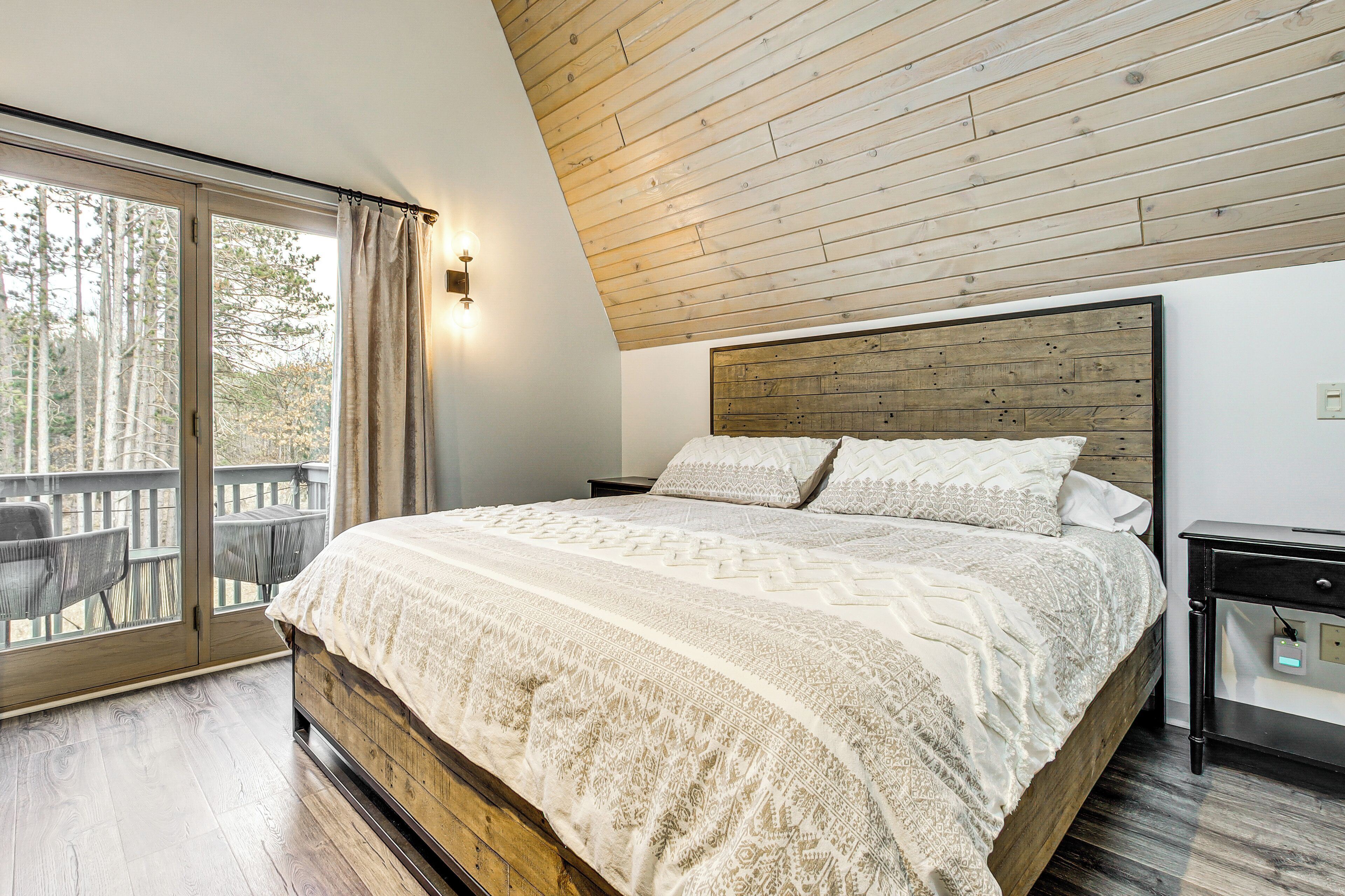 A bedroom with a wooden headboard, a large bed with patterned bedding, a nightstand, and a view through a sliding glass door to an outdoor deck with chairs.