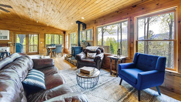 A cozy cabin interior with a wooden ceiling, large windows, a leather sofa, a blue armchair, and a stone fireplace.
