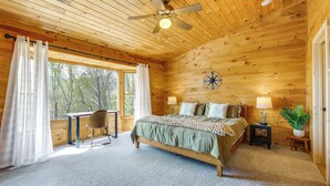 A wooden cabin bedroom with a bed, a desk, a chair, a ceiling fan, and a window with curtains.