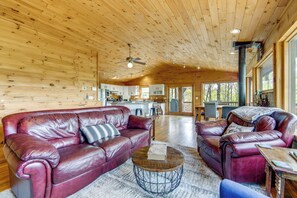 A spacious living area with a large leather sofa, a coffee table, and a dining table. The room features wooden walls and ceiling, a kitchen area in the background, and large windows allowing natural light. - 10 Mi to Waynesville: Cabin w/ Hot Tub & Mtn Views (Balsam)