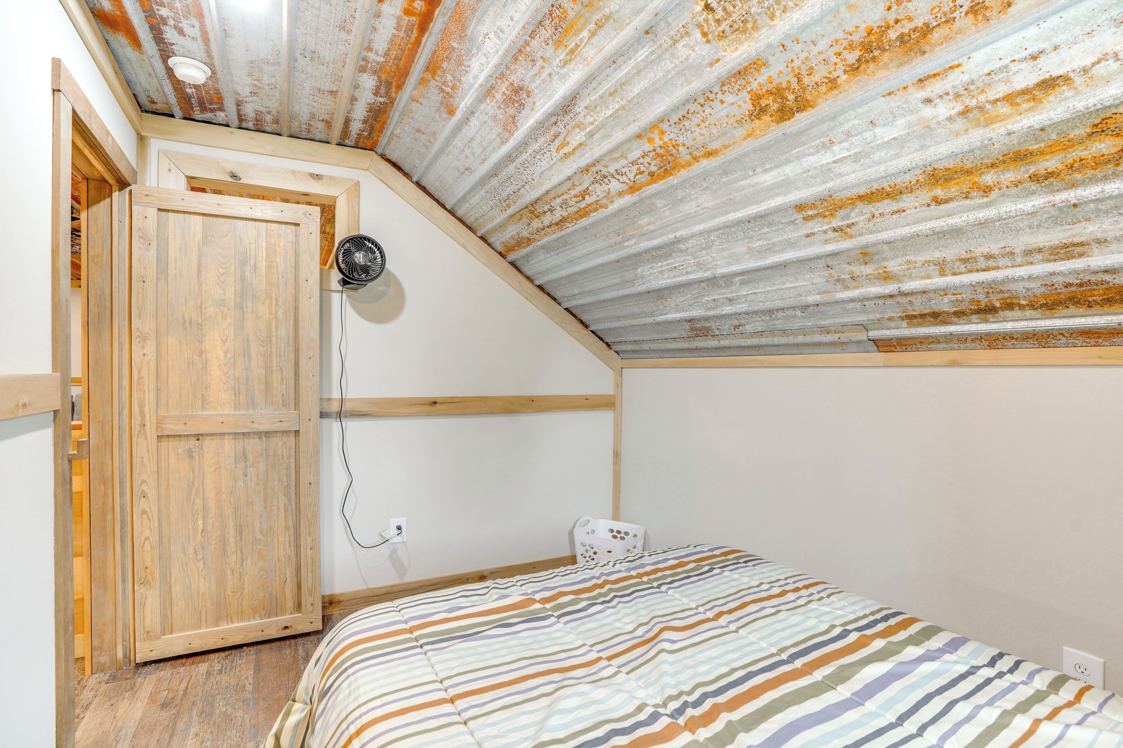 A bedroom with a sloped ceiling, a wooden door, a bed with striped bedding, and a wall-mounted fan.
