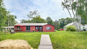Une maison rouge avec une terrasse en bois menant à la porte d’entrée, entourée d’une cour verdoyante et d’arbres.
