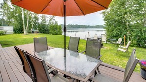 Une table avec un plateau en verre, quatre chaises, un parasol orange et une vue sur un lac.