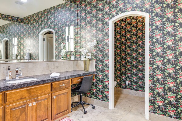 Un baño con papel tapiz floral, armarios de madera, encimera de granito y una silla de escritorio negra.