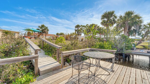 A wooden deck with a round table and chairs, surrounded by lush greenery and palm trees. - Canal-front Home w/ Private Dock in North Port! (North Port)
