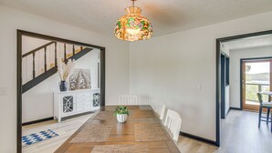 Comedor con mesa de madera, sillas blancas, una lámpara colgante decorativa y un mueble blanco con cajones.