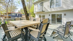 A deck with a wooden table and chairs, a swing chair, and a house with a balcony in the background. - Balcony & Fire Pit: Home on Lake of Ozarks! (Roach)