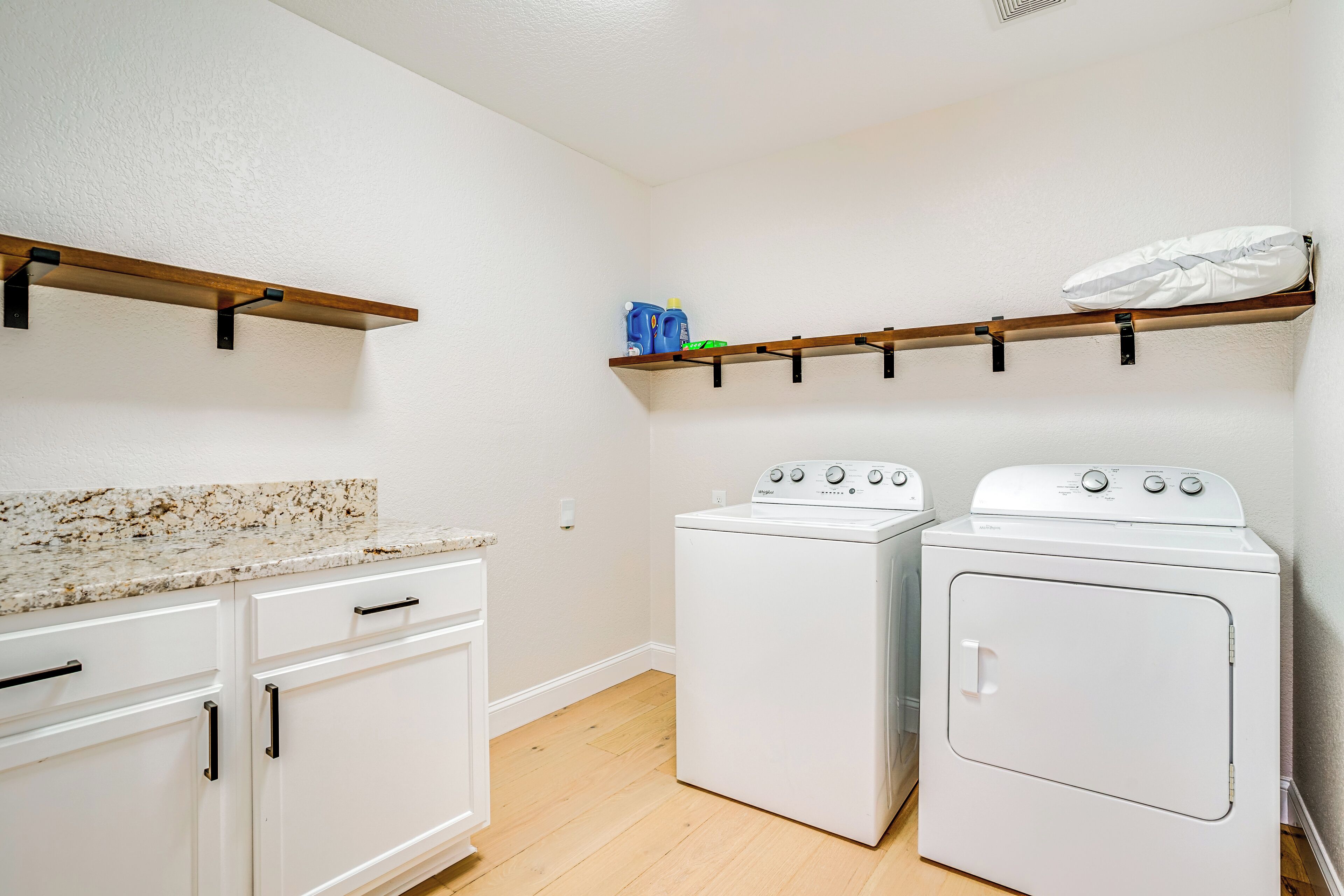 Un espace buanderie équipé d’un lave-linge et d’un sèche-linge, avec des étagères en bois au-dessus et des meubles blancs dotés de poignées noires.