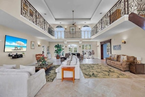 A spacious living room with a high ceiling, a balcony with ornate railings, and a large window providing natural light.