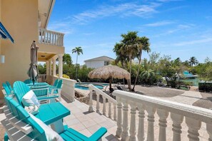 A balcony with turquoise chairs and a thatched umbrella, overlooking a pool and palm trees.