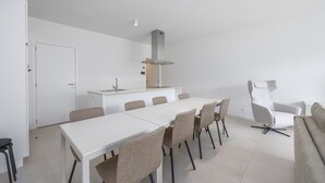 Un espace repas moderne et minimaliste, avec une table et des chaises blanches, une cuisine intégrée dotée d’une hotte en acier inoxydable et un fauteuil à revêtement gris.