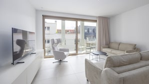 Un salon moderne avec un téléviseur à écran plat, un canapé gris, un fauteuil gris, une petite table ronde et un balcon avec une porte vitrée.