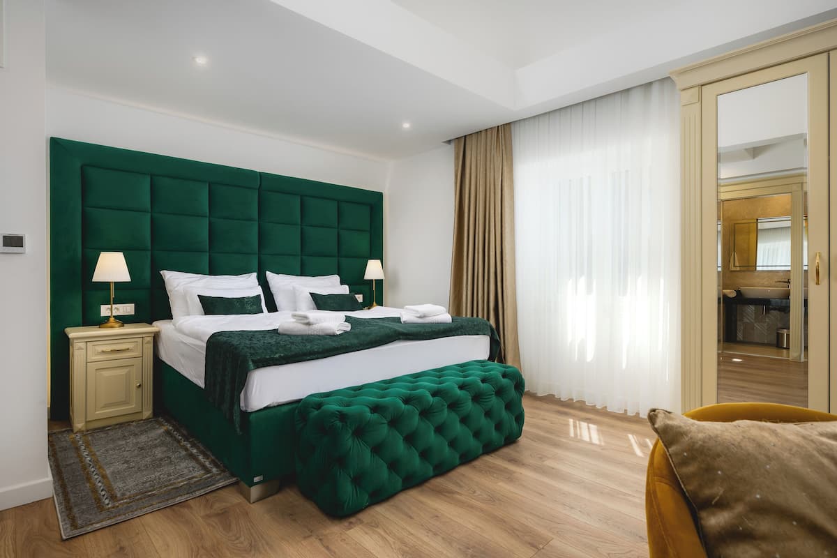 A hotel room with a green upholstered bed, a bedside table with a lamp, a wooden floor, and a view of the bathroom through a glass door.