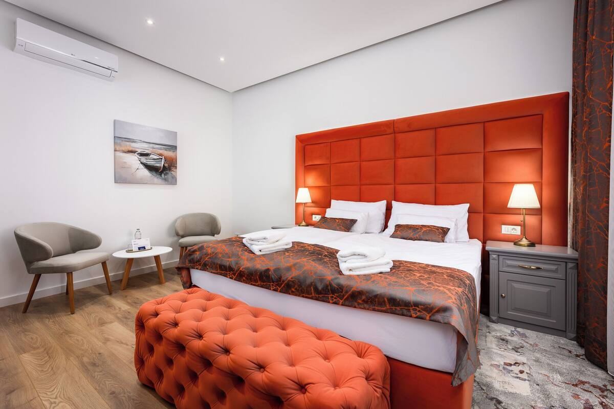 A modern hotel room with a large bed, a tufted orange headboard, two bedside lamps, a small table with a bottle, and a chair.