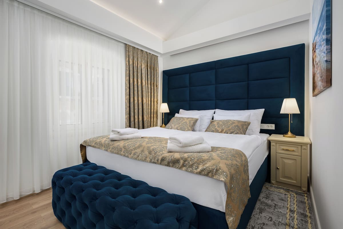 A modern hotel room with a large bed, a tufted bench, a nightstand with a lamp, and a painting on the wall.