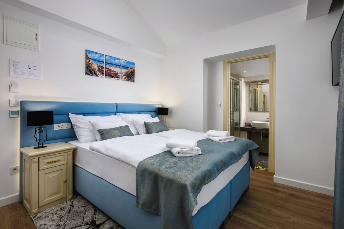 A modern bedroom with a blue bed, white bedding, a nightstand with a lamp, and a painting on the wall.