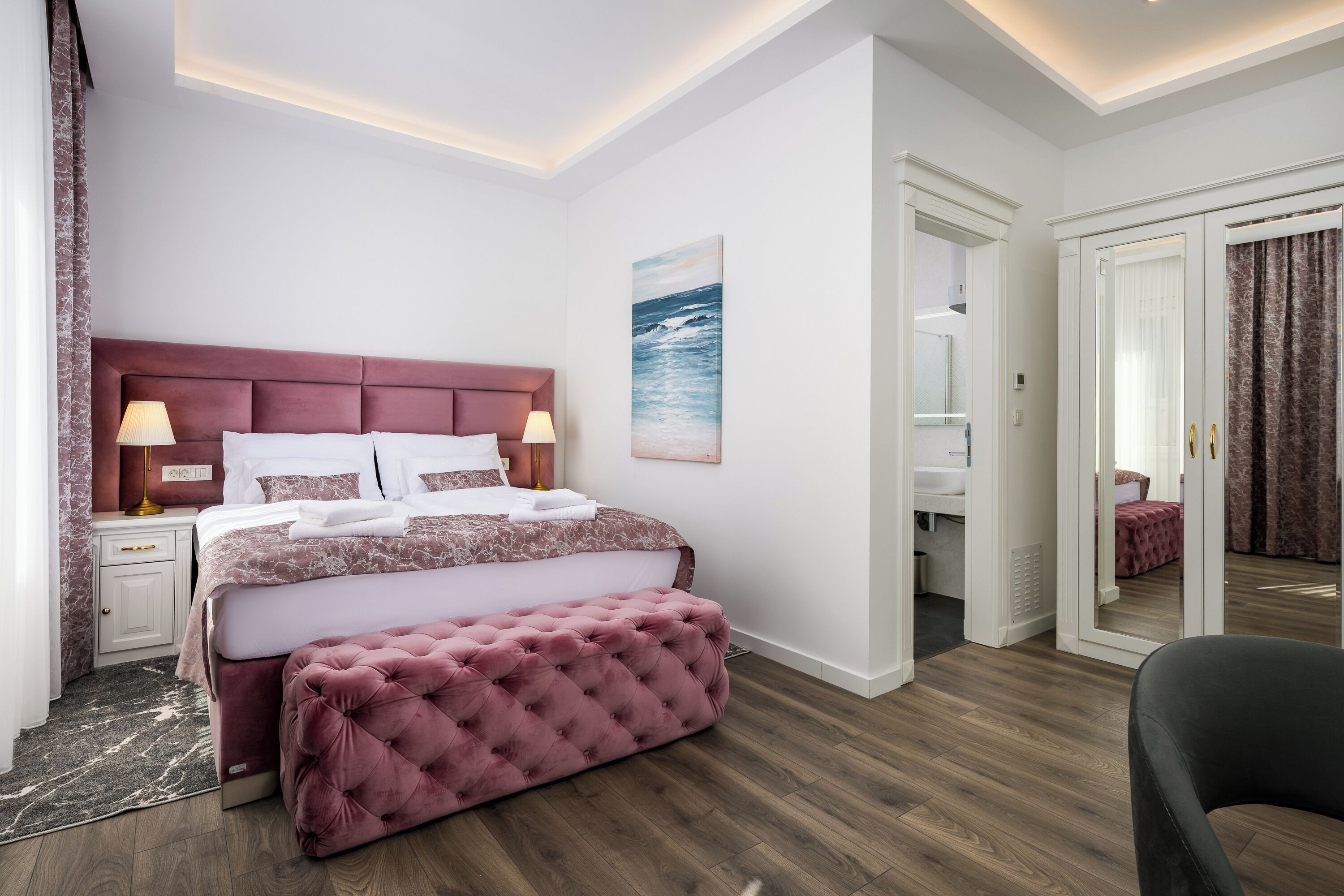A modern bedroom with a large bed, bedside lamps, a tufted bench, a painting, and a view into an adjacent room.