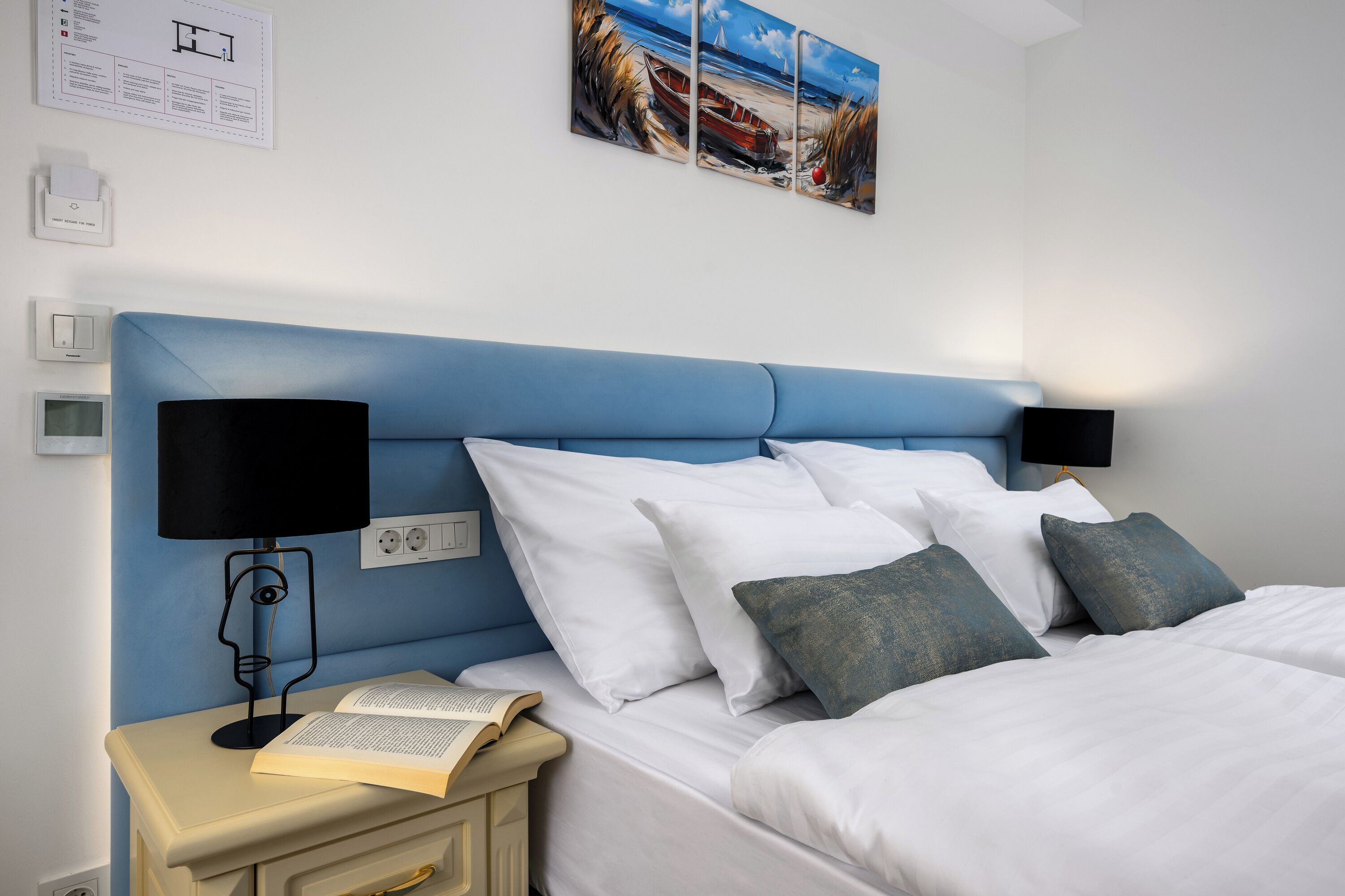 A neatly made bed with white and grey pillows, a bedside table with a book and a lamp, and a wall-mounted painting of a beach scene.