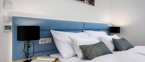 A neatly made bed with white and grey pillows, a bedside table with a book and a lamp, and a wall-mounted painting of a beach scene.