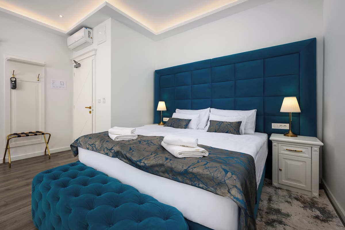 A modern hotel room with a large bed, a blue headboard, two bedside tables with lamps, a white door, and a small bench.