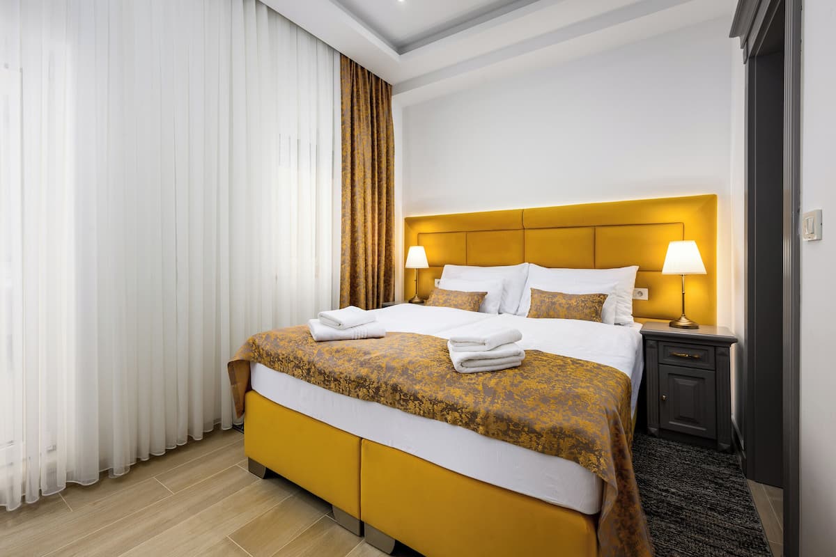 A hotel room with a large bed, two bedside lamps, a dark nightstand, and a large window with white curtains.