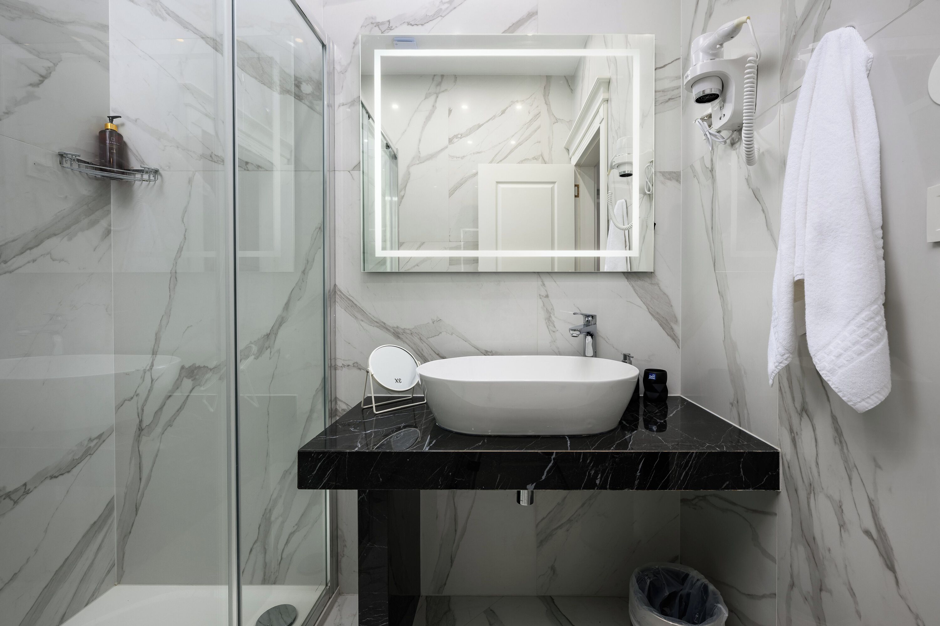 Een moderne badkamer met een marmeren wand, een zwart aanrecht, een wit wastafel, een spiegel, een haardroger en een handdoekhouder.