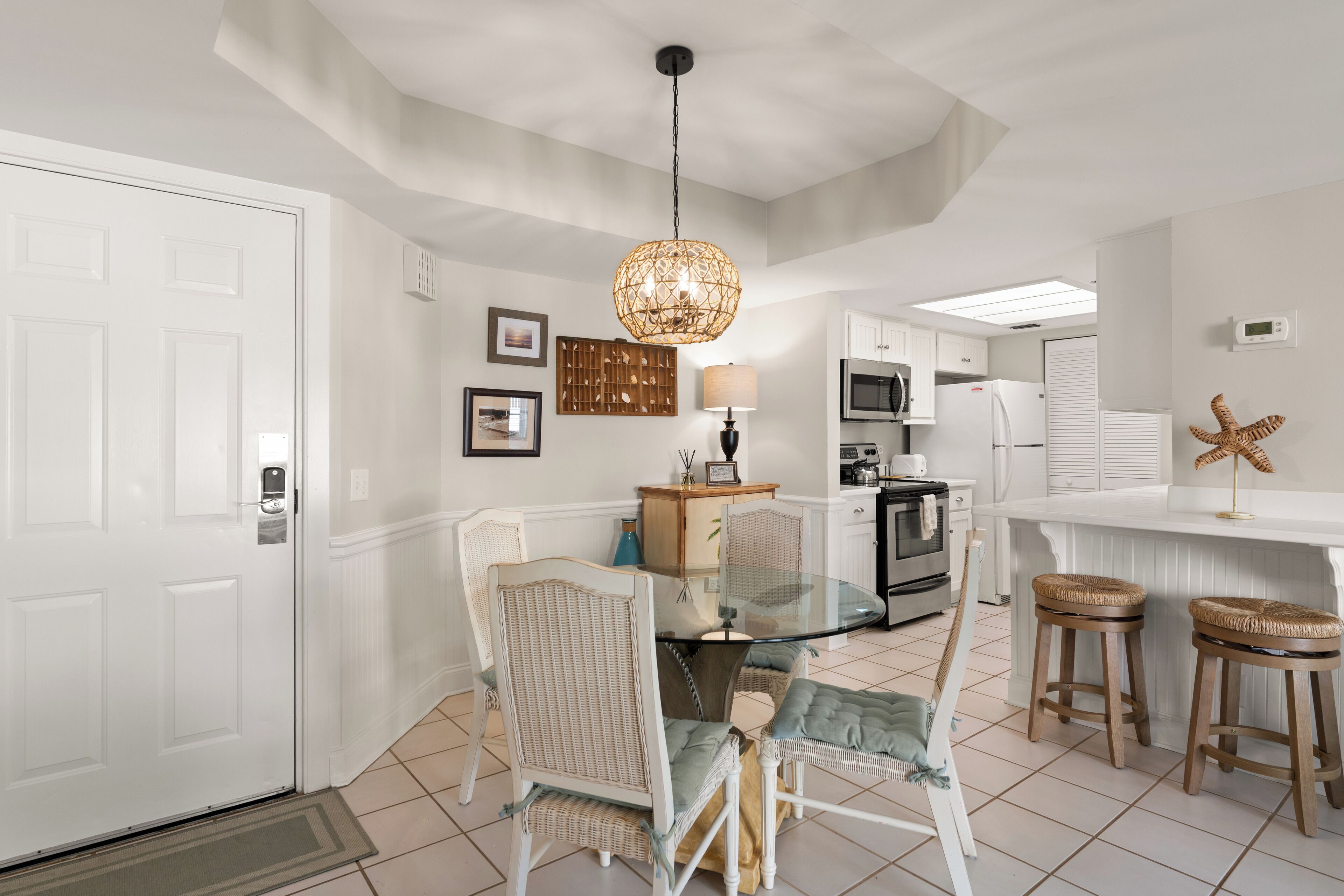 A modern kitchen with white cabinetry, a glass dining table, wicker chairs, and a hanging light fixture.