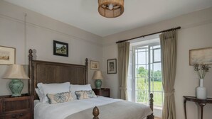 A bedroom with a large bed, wooden headboard, two bedside tables with lamps, a window with curtains, and framed pictures on the walls.