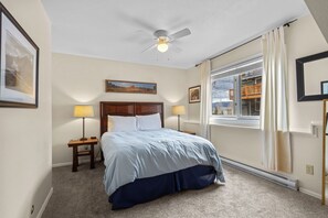 Un dormitorio con una cama, dos mesitas de noche con lámparas, un ventilador de techo y un ventanal con cortinas.