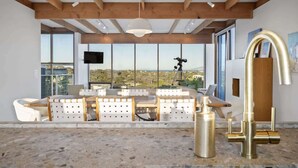 Une cuisine moderne dotée d’un grand îlot central, d’un plafond haut avec des poutres apparentes et de grandes fenêtres offrant une vue sur le paysage.