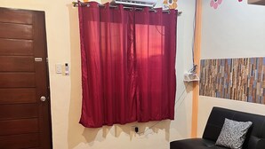 A room with a wooden floor, a red curtain, a black sofa with a patterned pillow, and a wooden door.