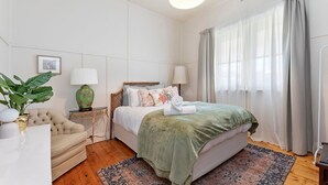 Quarto bem iluminado com cama, mesas de cabeceira, uma cadeira e uma janela grande com cortinas.