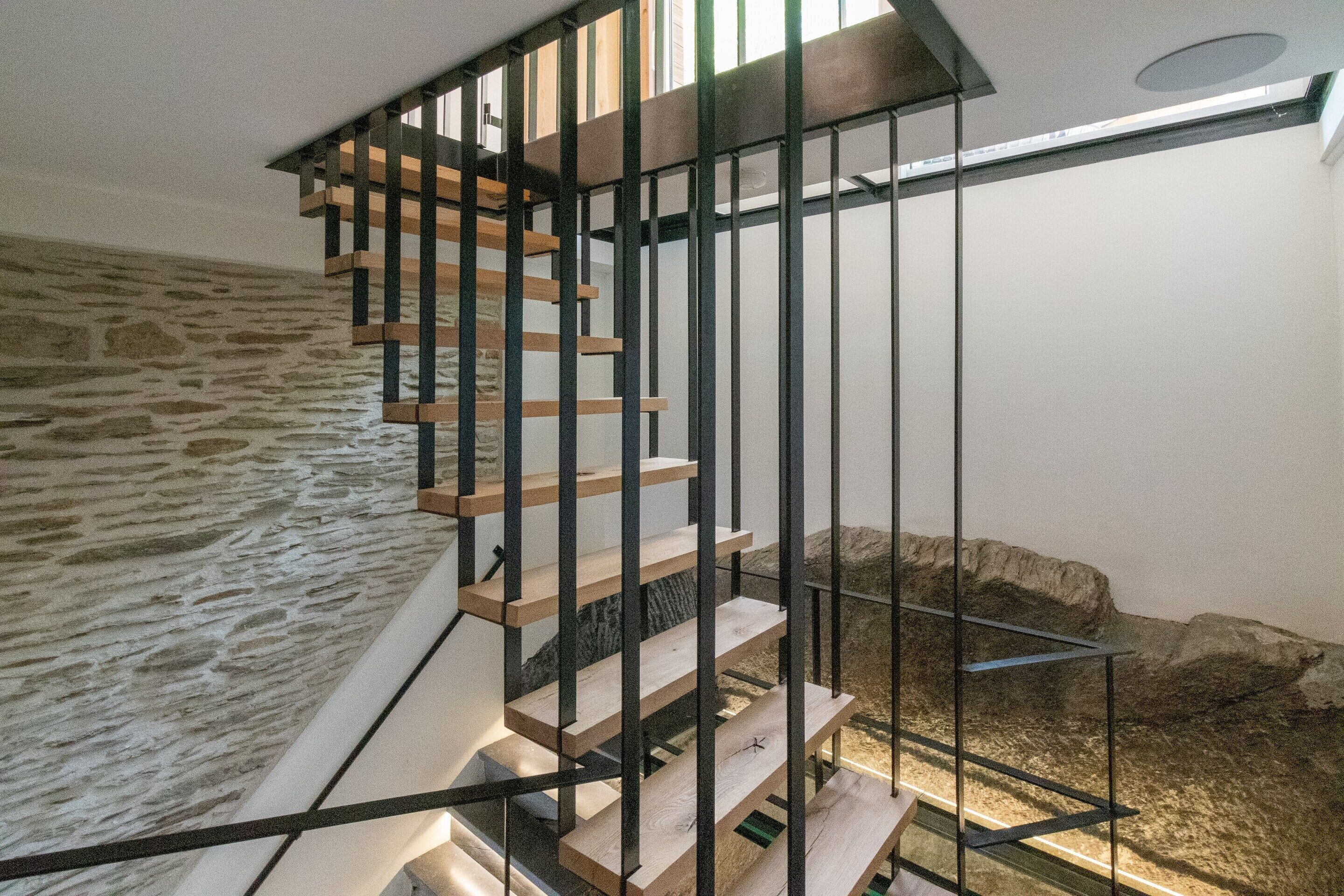 A staircase with wooden steps and black metal railings, leading to a landing with a stone wall and a window.