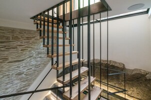 Una escalera de madera con barandillas de metal negro, que conduce a un rellano con un muro de piedra y una ventana.