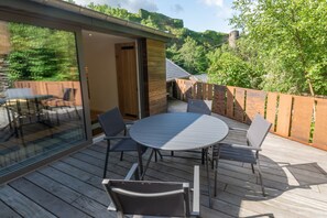 Une terrasse en bois avec une table ronde et des chaises, une porte vitrée donnant sur une pièce, et une vue sur une colline boisée avec, au loin, une structure ressemblant à un château.