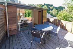 A wooden deck with a glass door leading to a room, a round table with chairs, and a view of a forested area. - Holiday Home in La Roche With Castle Views (La Roche-en-Ardenne)