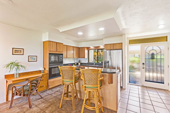 A kitchen with wooden cabinets, a dining area with two wicker chairs, and a large island. - 226-4 Steps to Surf and Sun (Lahaina)
