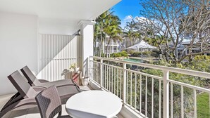 A balcony with wicker furniture, a white table, and a view of a pool and buildings.
