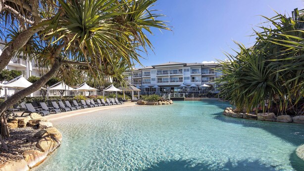 A clear pool with lounge chairs and umbrellas, surrounded by palm trees and a building with multiple balconies.