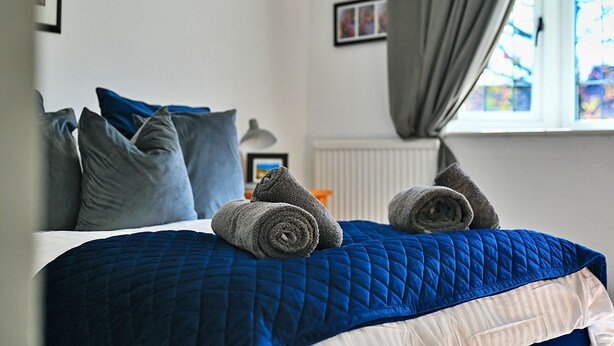 A neatly made bed with a blue quilt, grey pillows, and rolled towels.