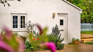A white building with two windows and a door, surrounded by greenery and a wooden deck. - The Nook (Goulceby)
