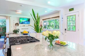 A kitchen with a gas stove, a bowl of fruit, a vase with flowers, and a window with a view of greenery.