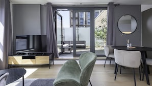 A modern living room with a flat-screen TV, a dining table with chairs, a sofa, and a large window offering a view of the outside.