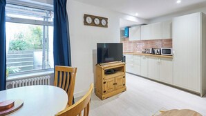 A compact living space with a dining area, a TV on a wooden stand, a kitchen with white cabinets, and a view through a window.