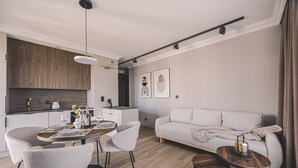 A modern living room with a sofa, dining table, and chairs. There is a kitchen area with a wooden cabinet and a hanging light fixture. - Industria Apartment Zawisza Parking Balkon (Bydgoszcz)
