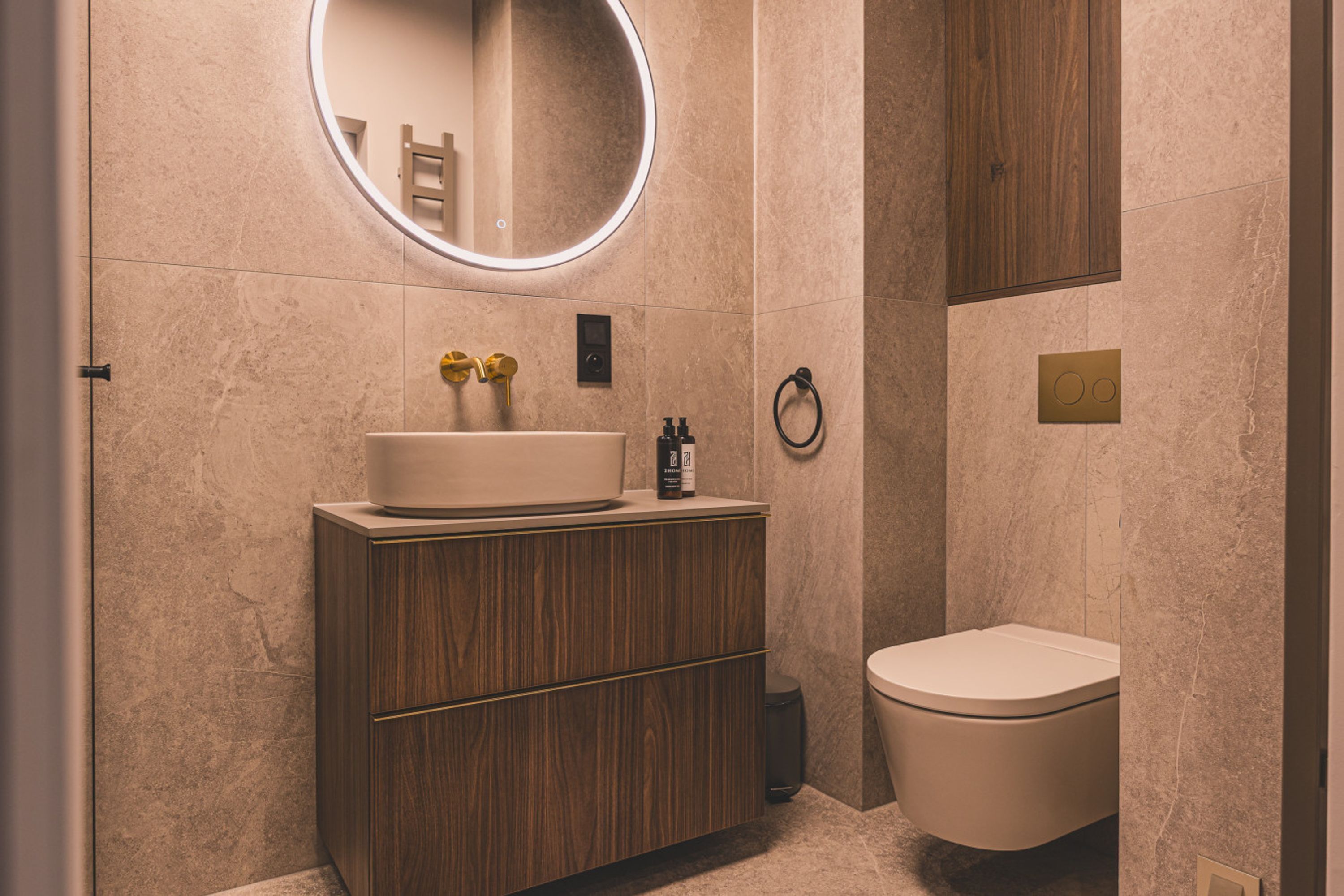 A modern bathroom with a wooden vanity, a round mirror, a toilet, and a towel ring.