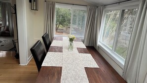 Ein Esszimmer mit einem langen Tisch, Stühlen, einer Vase mit Blumen und großen Fenstern mit Vorhängen.