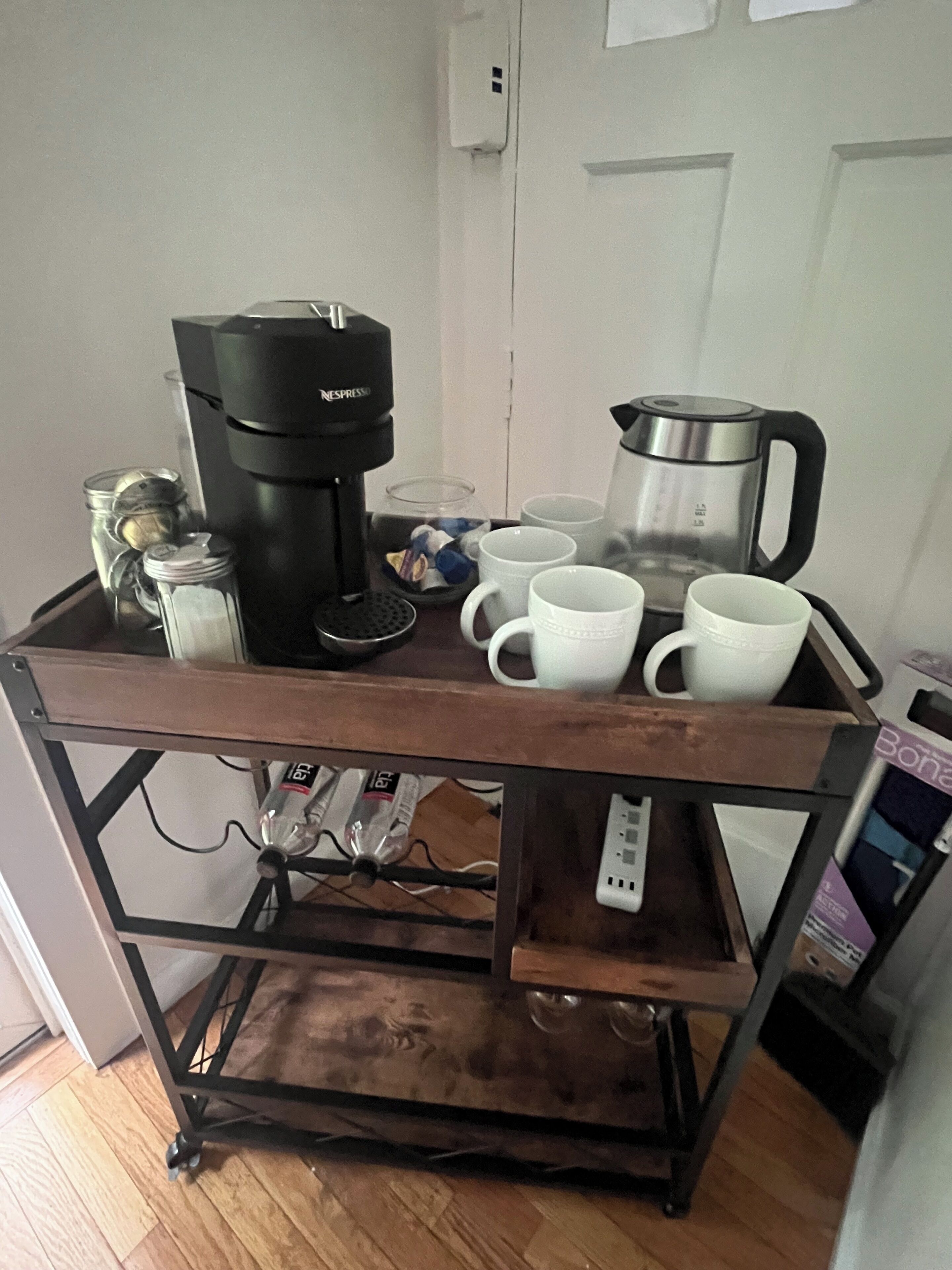 A coffee station with a Nespresso machine, a glass carafe, and several white cups on a wooden cart.